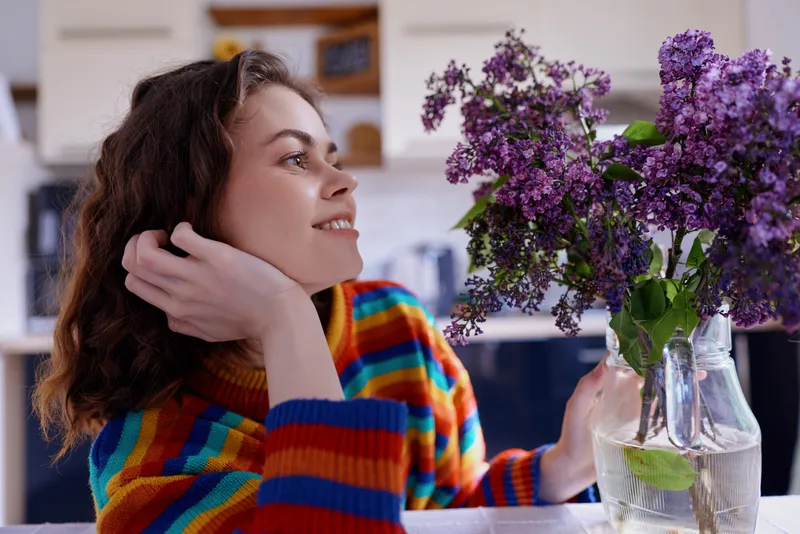 Smiling woman with curly hair sits in a bright kitchen admiring purple lilac flowers in a glass vase. Cozy home lifestyle moment, warm light, calm mood, everyday comfort.