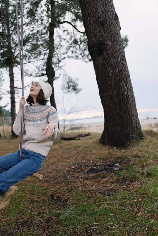 Swing scene with a woman in a cozy sweater and beanie on a rope seat, surrounded by trees and grass near a calm water edge, capturing carefree outdoor leisure and connection with nature.