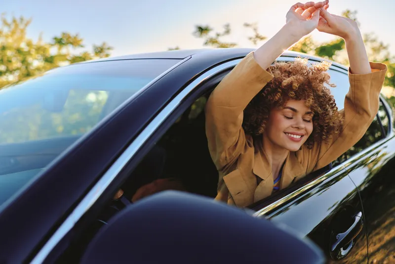 Woman leaning out of a sleek car, enjoying a carefree outdoor moment, warm sunlight, breezy park setting, retro film color, vibrant natural tones, candid lifestyle vibe