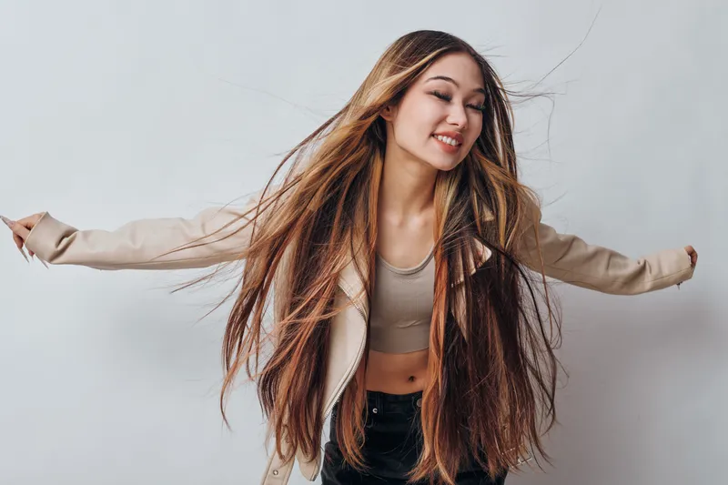 Young woman with long hair wearing beige jacket and crop top smiling with eyes closed on gray background for beauty and joyful lifestyle portrait.