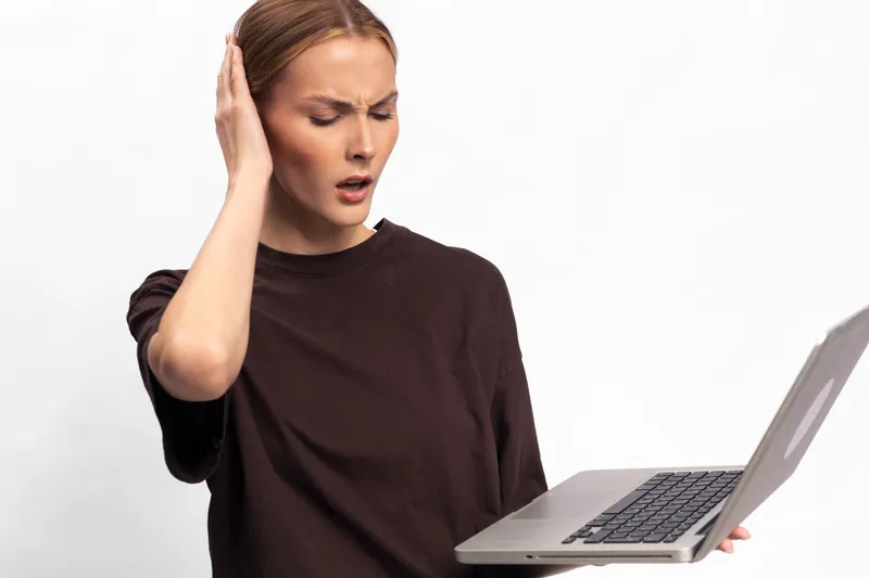 Confused woman with laptop holds head with hand, looking frustrated and surprised. Isolated on white background. Concept of technology problems, stress, and digital frustration.