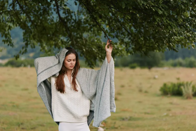 Cheerful woman outdoors, draped blanket and sweater, arms raised in a relaxed lifestyle moment, fresh air, soft breeze, natural colors, green meadow background in view