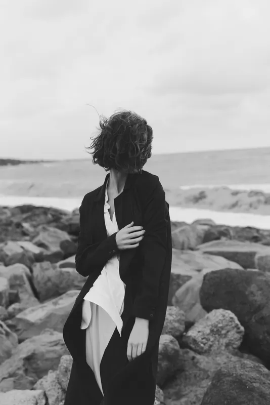 Woman in a long coat on a rocky shore, wind tousling hair, monochrome scene with sea spray, coastal mood, fashion style, solitary silhouette near the horizon.