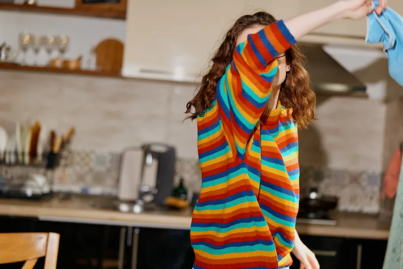 Vibrant young woman wearing a colorful striped sweater dances barefoot indoors in a bright kitchen, her lively movement and cheerful expression filling the cozy space with energy and joy