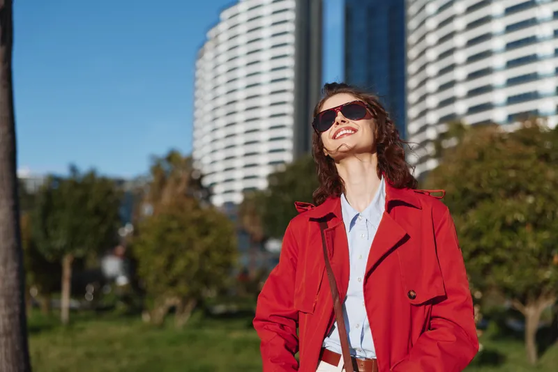Young woman in a red coat and sunglasses smiles confidently outdoors in a sunny urban park with modern buildings in the background.