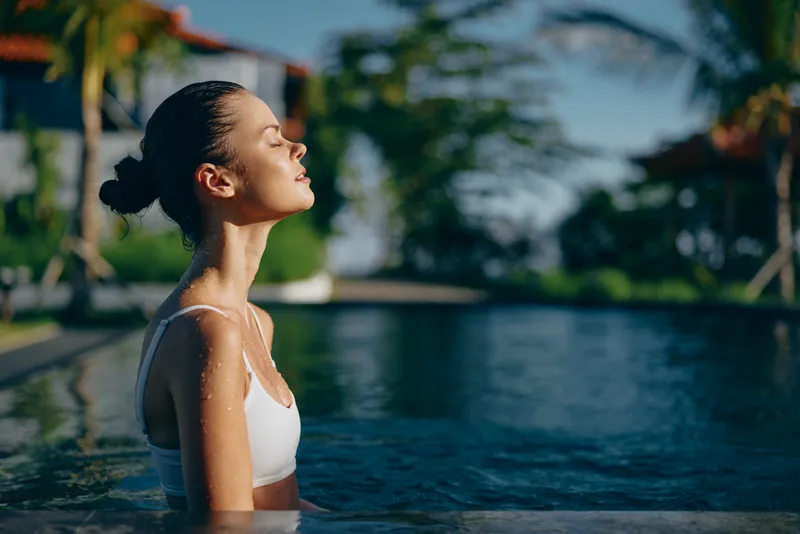 A young woman relaxes in a sunlit pool, eyes closed, savoring a peaceful moment as warm water surrounds her in a bright outdoor setting, conveying calm and mindfulness.