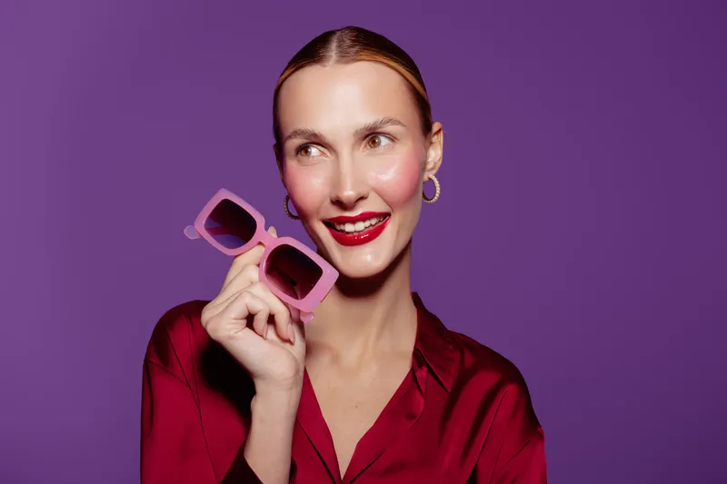 Smiling woman in red blouse holds pink sunglasses near face on purple background. Concept of fashion, beauty, accessories, and stylish portrait with bright makeup and confident expression.