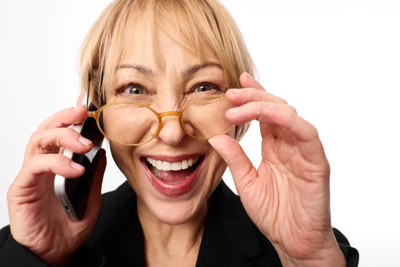 Blonde older woman with glasses smiling while holding phone to ear, excited expression, close up portrait on white background, modern eyewear, friendly communication call, laughing