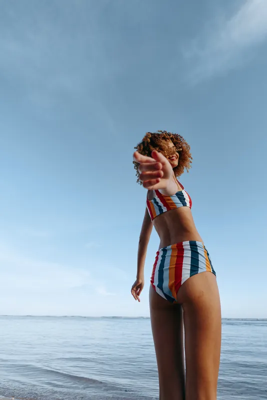Young woman in striped swimsuit stands at beach by ocean under clear summer sky turning to camera holding curly hair outdoors sunlit shoreline calm waves minimal composition background