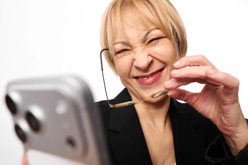 Older woman holding spoon near smartphone, eyes closed and smile, close up portrait in casual home setting, soft studio light on white background, eating moment
