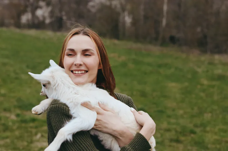Cheerful woman outdoors, smiling while cuddling a white goat kid, sunlit field scene, relaxed posture, soft breeze, natural colors, rustic countryside atmosphere, joyful moment