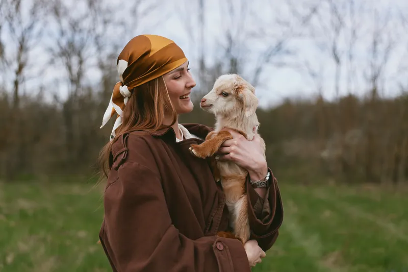 Smiling woman in a brown coat holds a young goat kid in a grassy field, capturing a warm lifestyle moment with animals, nature, and gentle companionship, showcasing simple happiness outdoors and