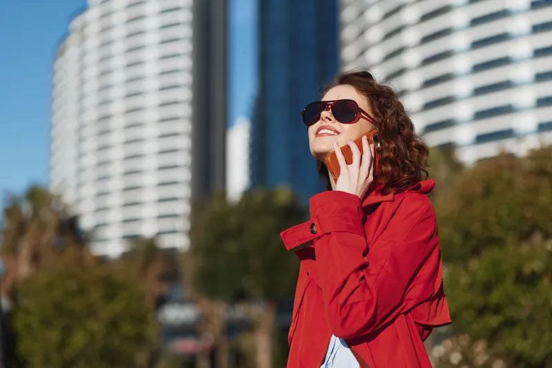 Young woman in a red coat smiles while talking on a smartphone outdoors in an urban setting on a sunny day with modern buildings in the background.