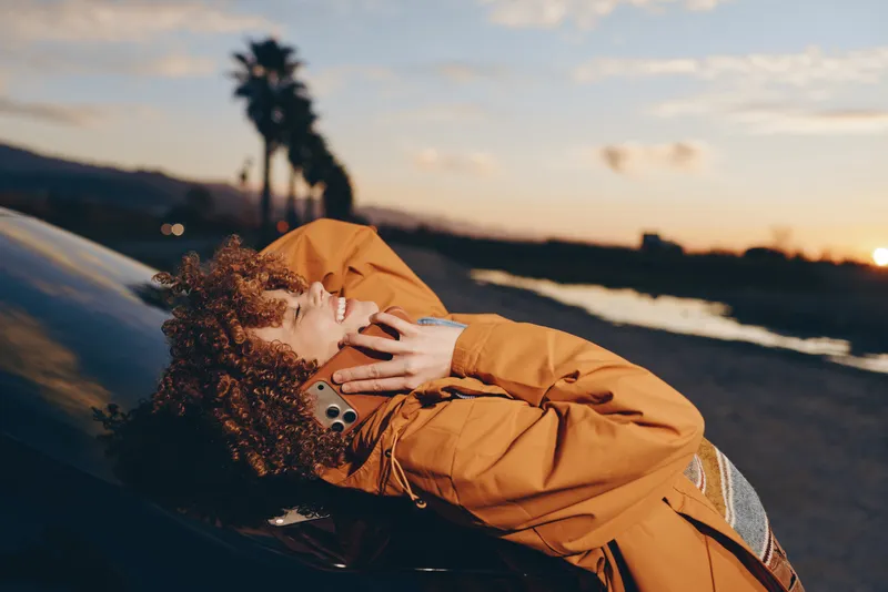 Woman lifestyle portrait by the ocean coast, outdoor scene at sunset, curly hair in wind, wearing an orange coat, beside a car on a coastal road, warm golden light, calm mood