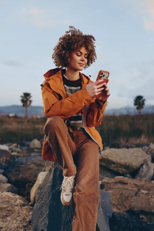 Woman lifestyle portrait outdoors, curly hair, orange jacket, casual jeans, holding smartphone while sitting on rocks, sunny nature with palm trees, travel leisure concept