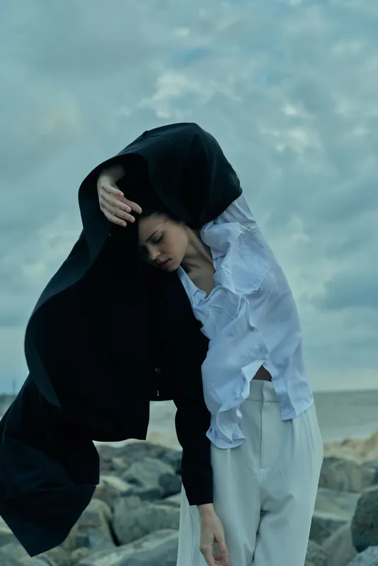 Woman in flowing shirt and hijab on rocky coastline, wind blowing fabric, moody ocean backdrop, modest fashion, dramatic natural light, serene coastal style