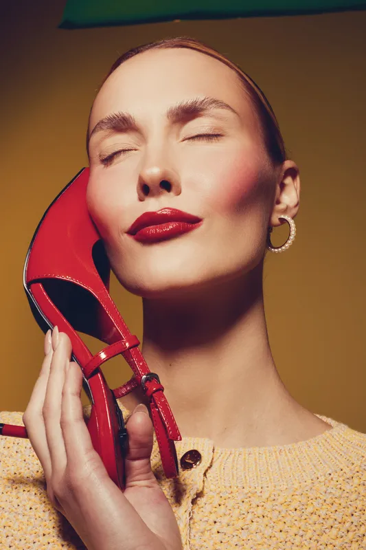 Close-up of a woman with red lipstick holding a red high heel shoe near her face. Yellow background and soft lighting create a stylish and elegant fashion concept.