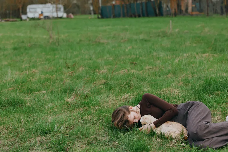 Calm woman lying on grass, hugging a goat kid in an outdoor scene. Lifestyle image highlighting connection with animals, comfort, calmness, and wellbeing. Soft natural light creates a warm, intimate