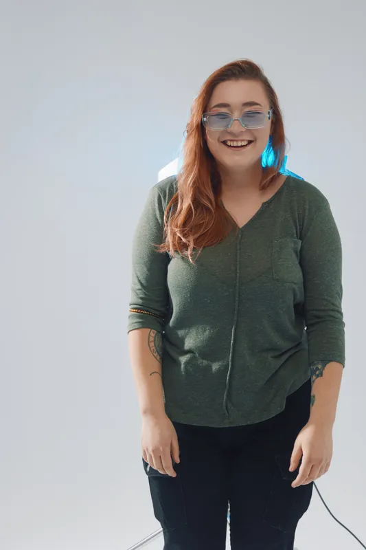 Smiling woman with long red hair wearing glasses and a green sweater. She stands relaxed against a plain background. Concept for beauty fashion and casual style.