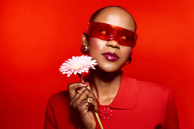 Elegant black woman with dark skin wearing red sunglasses and a red outfit holds a pink flower against a bright red background. Concept of fashion, beauty, and confident style.