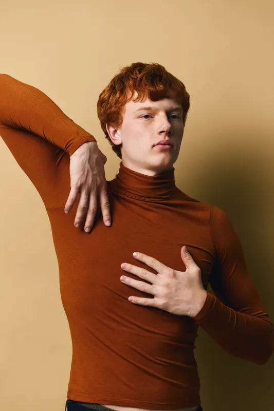 Young man with red hair posing in rust color turtleneck sweater against beige background. Fashion model with pale skin and serious expression in studio portrait photography.