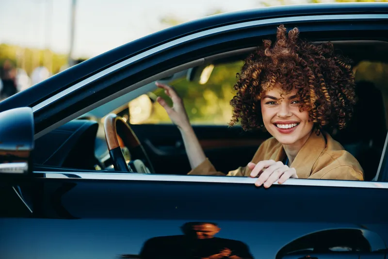 Smiling woman with curly hair sits in a car, hands on the steering wheel. Casual lifestyle shot with daytime lighting, warm retro film color tones and a candid, friendly mood.