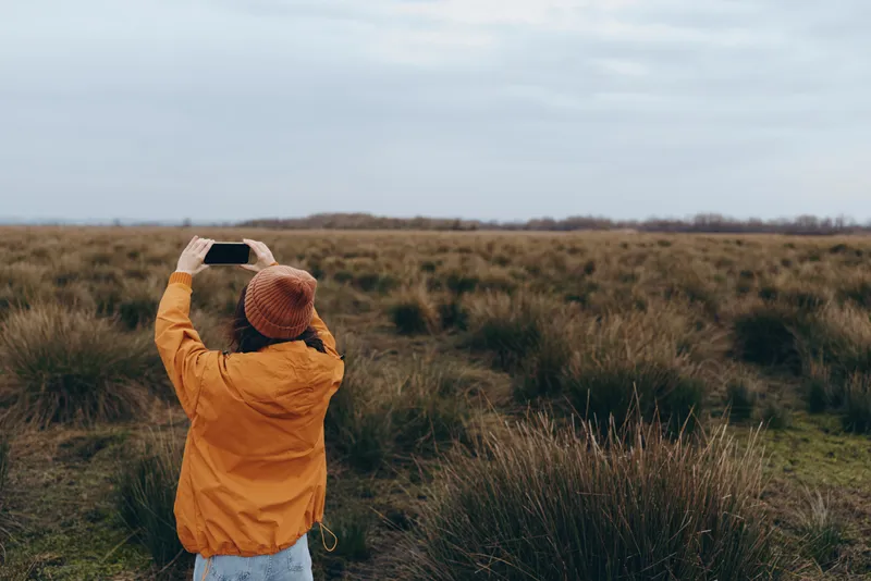 Woman in orange jacket and hat standing outdoors in a grassy field, facing horizon and cloudy sky, calm winter lifestyle mood, back view, wide landscape scene, for solitude and fresh air