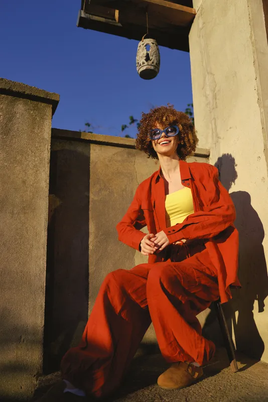 A stylish woman in a bright red suit sits outdoors, wearing sunglasses and a confident smile. Sunny rooftop setting conveys bold fashion and casual lifestyle. Light brightens scene