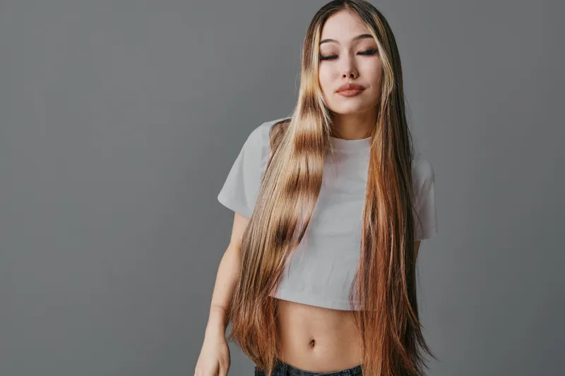 Young woman with long straight hair posing with closed eyes in casual outfit against grey background, showing calmness and natural beauty in studio portrait.