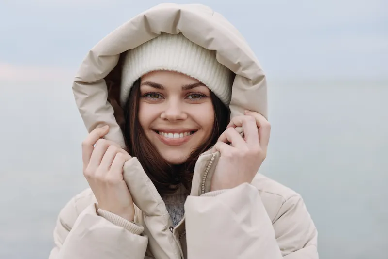 Woman in a warm hooded coat smiles outdoors by the water, winter fashion and cozy outerwear for cold weather, serene seaside setting.