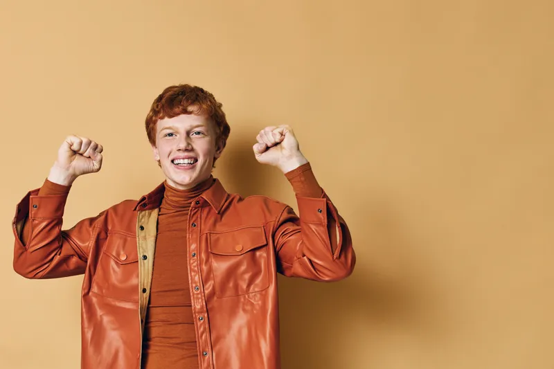 Happy young man with red hair and casual outfit celebrating success with raised fists, smiling and standing against beige background in studio portrait.