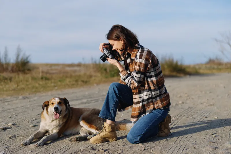 Photographer kneels on dusty ground, capturing a playful dog in an open outdoor landscape, wearing a plaid shirt and jeans, camera ready under a bright sky and sparse vegetation.