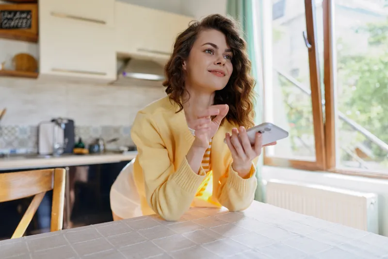 Smiling woman in a yellow cardigan sits at a kitchen table using a smartphone, enjoying a relaxed home moment. Bright daylight and natural light create a cozy atmosphere, casual lifestyle.