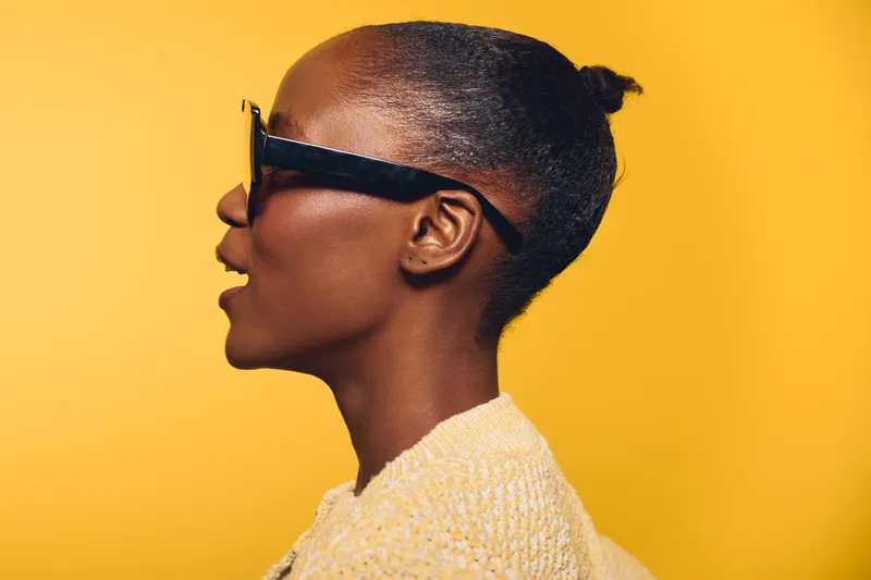 Profile of an African woman with dark skin wearing black sunglasses and a light sweater against a yellow background. Concept of fashion, style, and confident attitude.
