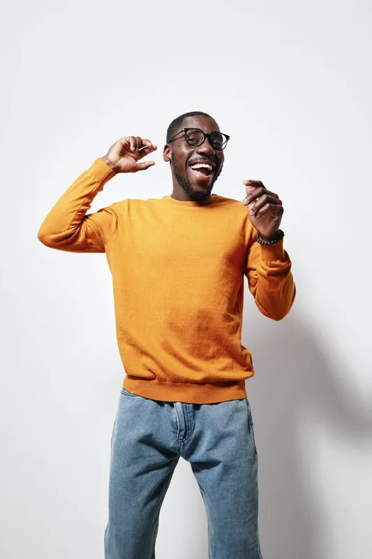 African American man with dark skin wearing orange sweater and glasses smiling and dancing against white background in casual jeans and wristwatch.