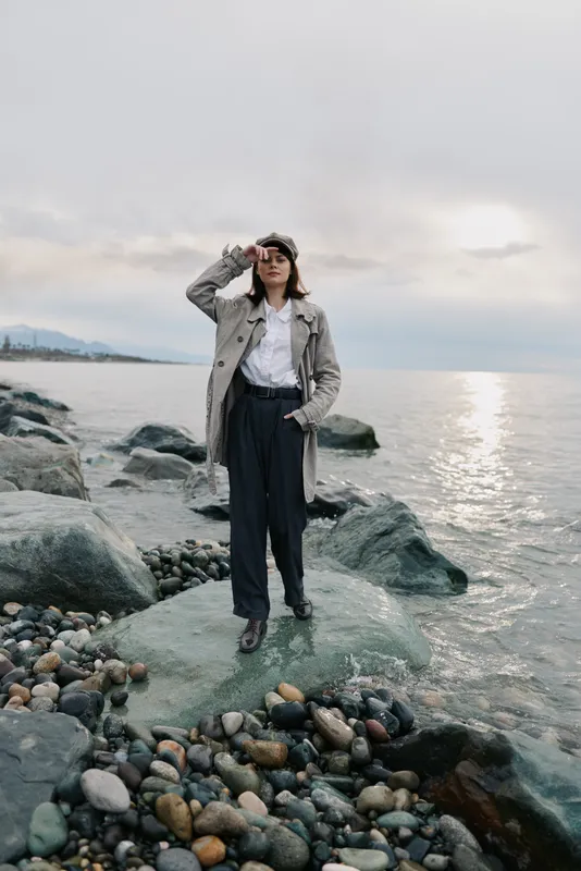 Woman in trench coat stands on rocky shore near calm sea, touching forehead. Overcast sky and soft sunlight create a reflective mood in this outdoor coastal scene.