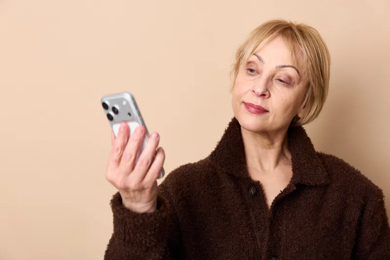 Elderly woman portrait indoors holding a smartphone, relaxed posture, soft natural light, warm beige background, gentle smile, focused expression, technology communication moment