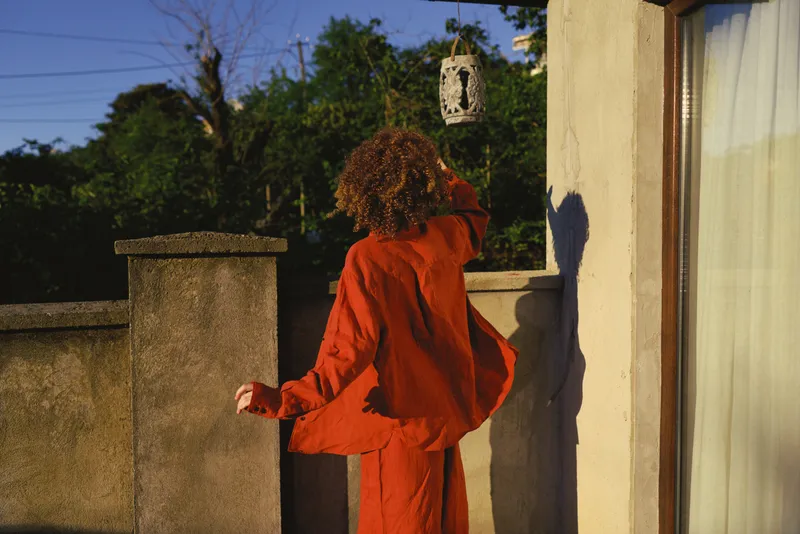 A young woman in a bright red jumpsuit stands on a balcony, looking back over her shoulder as a birdhouse hangs nearby