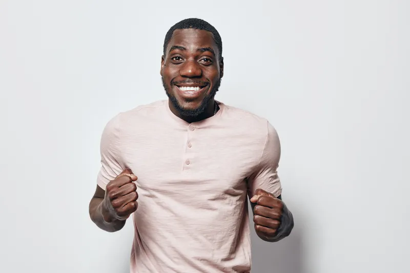 African American man with dark skin smiling and showing excitement with clenched fists wearing light pink shirt on white background. Happy male portrait with positive emotion and casual style.