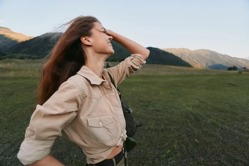 Young woman with long brown hair enjoys fresh air and freedom while standing in a wide open field with mountains in the background during daytime.