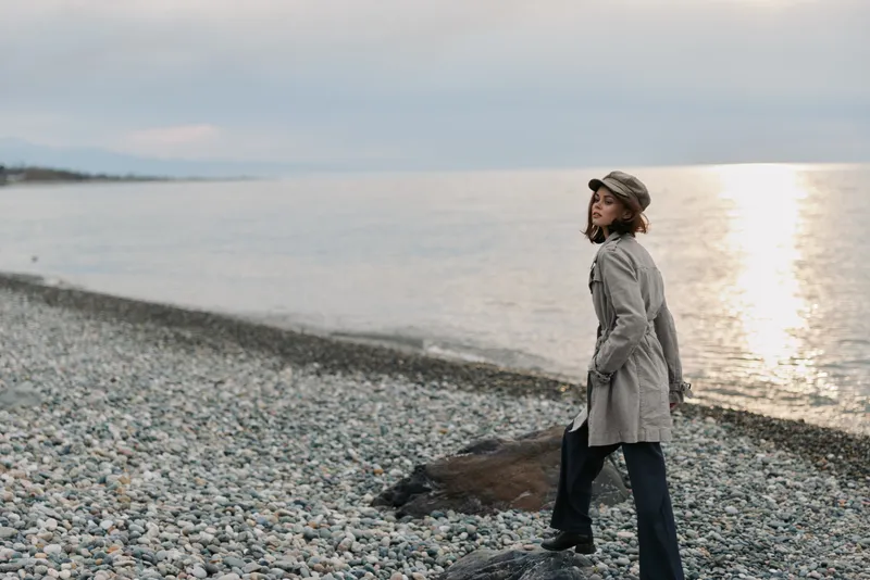 Woman in a gray coat and cap stands on a pebble beach looking over the sea during sunset. The calm water and cloudy sky create a peaceful and contemplative mood.
