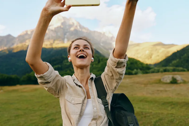 Young woman takes a selfie while hiking in nature, smiling at the camera with mountains in the background, casual outfit, backpack, bright sunny day, lifestyle portrait, vibe