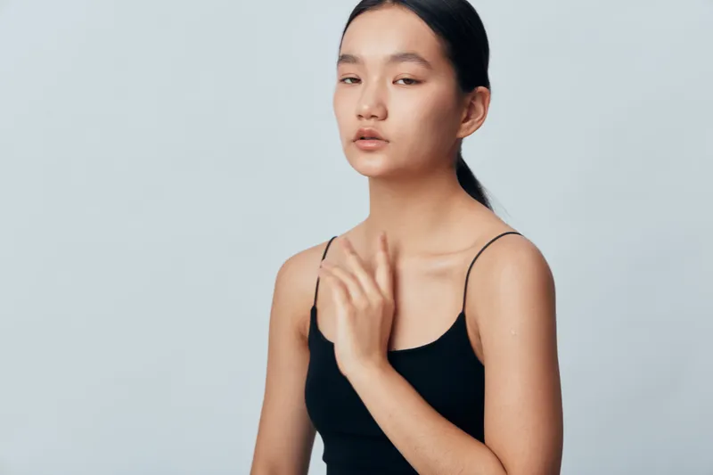 Young woman with smooth skin and natural face expression touching shoulder, wearing black sleeveless top, minimal makeup and simple hairstyle in studio setting with soft lighting.