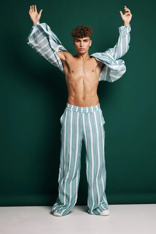 Young man with curly hair wearing striped pajama pants and shirt posing with arms raised against dark green backdrop for fashion and casual style concept with confident expression.