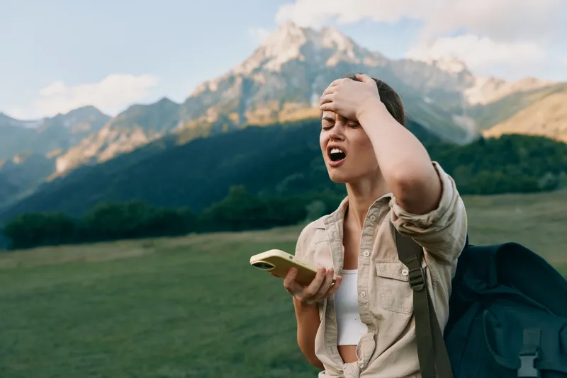 Young woman with backpack stands in a green meadow with mountains behind, checking her phone and wearing a casual shirt, a stressed expression on her face.