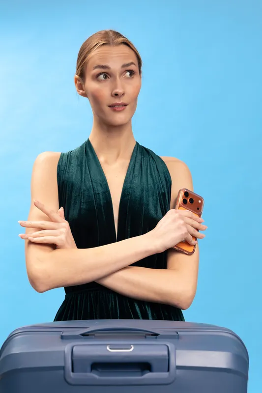 Young woman in elegant dark dress stands with crossed arms holding smartphone behind suitcase on blue background. Concept of travel, luggage, fashion, and thoughtful planning for trip preparation.