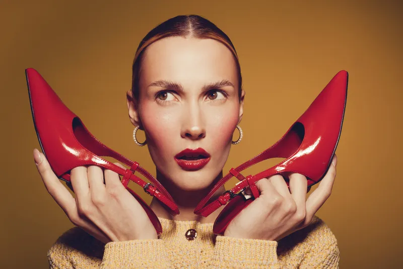 Young woman with red lipstick holds red high heel shoes near face, wearing beige sweater and hoop earrings. Studio portrait on brown background with fashion and beauty concept.