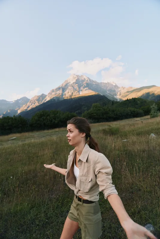 Young woman with long brown hair enjoys a scenic mountain landscape while standing in a grassy field during a clear day.