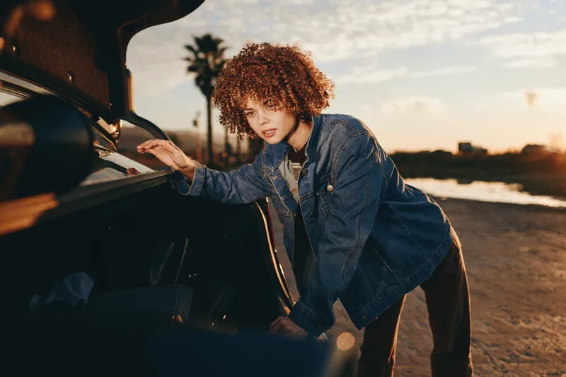 Woman lifestyle portrait, curly woman in denim jacket leans on open car door at roadside during sunset, golden hour sunlight, outdoors travel mood, casual trip, street road scene