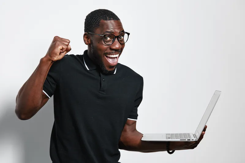 African American man with dark skin and glasses holding laptop and celebrating success with fist pump and smile on white background.
